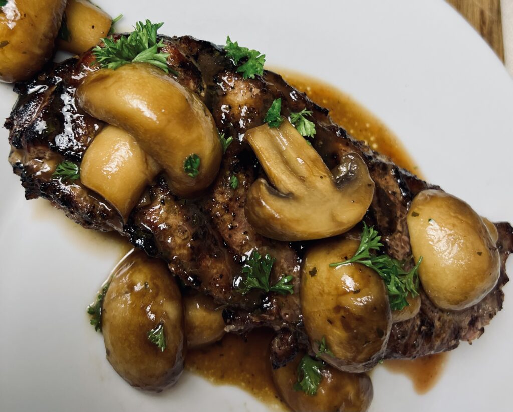 A juicy steak topped with golden-brown, buttery mushrooms, glistening with savory seasoning, creating the perfect steakhouse-style meal.