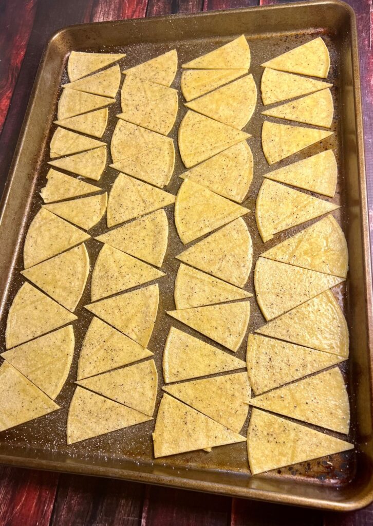 Tortilla chips arranged in a single layer on a baking sheet, lightly sprayed with oil and sprinkled with seasoning, ready to be baked to golden perfection.