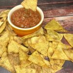 A close-up image of golden, crispy oven-baked tortilla chips arranged on a platter, paired with a vibrant red bowl of fresh salsa, garnished with cilantro.