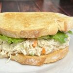 A close-up of a chicken salad sandwich on Italian bread. The sandwich is layered with creamy chicken salad, visible chunks of chicken, carrots, and celery, nestled between two slices of golden-brown Italian bread. Fresh lettuce leaves peek out from the edges, adding a touch of green. The bread has a soft texture with a slight crust, and the sandwich is served on a simple white plate. A few crumbs are scattered on the plate, suggesting freshness and a hearty meal.