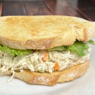 A close-up of a chicken salad sandwich on Italian bread. The sandwich is layered with creamy chicken salad, visible chunks of chicken, carrots, and celery, nestled between two slices of golden-brown Italian bread. Fresh lettuce leaves peek out from the edges, adding a touch of green. The bread has a soft texture with a slight crust, and the sandwich is served on a simple white plate. A few crumbs are scattered on the plate, suggesting freshness and a hearty meal.