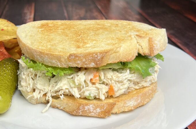 A close-up of a chicken salad sandwich on Italian bread. The sandwich is layered with creamy chicken salad, visible chunks of chicken, carrots, and celery, nestled between two slices of golden-brown Italian bread. Fresh lettuce leaves peek out from the edges, adding a touch of green. The bread has a soft texture with a slight crust, and the sandwich is served on a simple white plate. A few crumbs are scattered on the plate, suggesting freshness and a hearty meal.