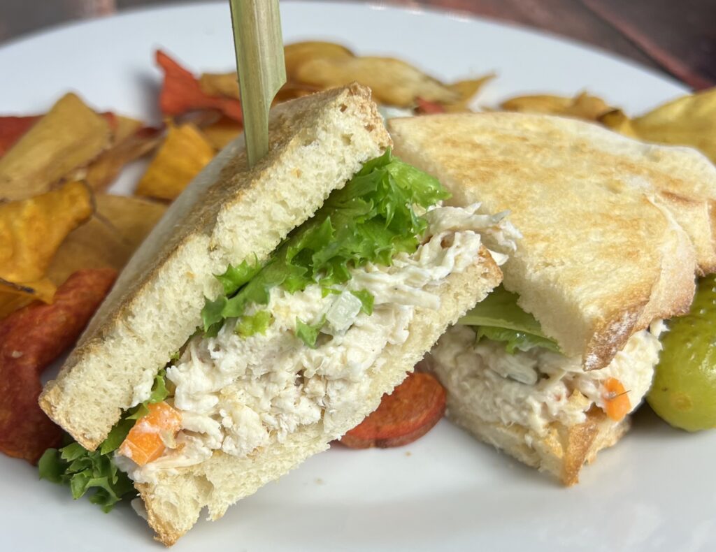 A chicken salad sandwich on Italian bread, cut in half to reveal its creamy filling, is served on a white plate. The sandwich is accompanied by a handful of colorful root chips and a crisp dill pickle spear on the side. The bread's golden crust and soft interior contrast with the creamy chicken salad and fresh green lettuce inside. The vibrant hues of the root chips and the pickle's bright green add a pop of color, creating an appetizing and well-balanced meal presentation.