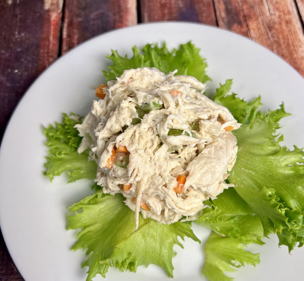 A scoop of creamy chicken salad sits on a fresh bed of vibrant green lettuce, served on a simple white plate. The chicken salad features chunks of tender chicken blended with a creamy dressing, lightly seasoned with Beau Monde seasoning and garlic salt. The crisp lettuce adds a fresh, colorful base that enhances the presentation of this classic dish.